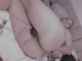 AmberDemons lying on a bed, legs up, wearing pink lace garter, purple dildo in her butt, black butt plug, blue bra, white towel, holding black remote, smiling.