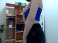 Steisii with light skin, blue tube top, and black jeans, covering her small tits with her hand, standing in a room with a wooden shelf and green wall.