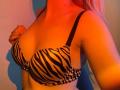 FlyFreedom has zebra-patterned tits in a black-strap bra, with her right hand near her chest. Her blonde hair is visible, and the photo has a red-orange light.