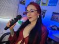 AmyStreetwise69 with red hair, glasses, and tattoos, holding a black microphone, wearing a red sheer top, showing her tits, smiling, sitting in a chair, blue light.