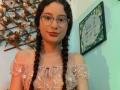 FluffydirtyMind with braided black hair, glasses, and pink floral off-shoulder top, showing her tits, has a cute smile and red lipstick, standing in a green-tinted room.