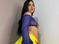 NaomyRosse with long black hair, blue sheer top, yellow skirt, and red lipstick, exposing her small tits and flat ass, standing against a plain wall.