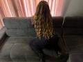 Niferttiti, with long wavy blonde hair, kneels on a dark gray couch, wearing black leggings and black boots, facing pink curtains, showing her curvy ass.