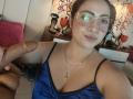 AlanaWest with glasses, blue satin top, holding a large, circumcised, erect penis. She has medium tits, light brown skin, and a slight smile. Background includes red pillows and decor.
