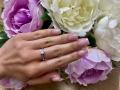 OliviaGrandas Hand mit rosafarbenen Nägeln, einem silbernen Ring und einem kleinen Diamanten, ruht auf weißen und rosa Pfingstrosenblüten. Ihre Finger sind leicht gespreizt..