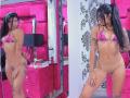 RoxyThunder with long black hair in a pink bikini top, standing naked with visible pussy, tattoos on right arm, in a pink and black room, smiling at the camera.