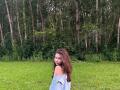 EmeraldCrivaro with long brown hair, looking back over shoulder, wearing an off-shoulder blue shirt, standing in green grass, forest of tall trees in background.