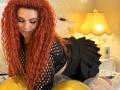 Niferttiti with red curly hair, black fishnet top, black skirt, on bed, looking down, surrounded by gold and white balloons, warm yellow lamp in background.