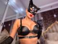 Mademoiselle Bella con maschera da gatto, reggiseno di pelle nera con fibbie e guanti, che mostra i suoi seni, le labbra rosse e il tatuaggio sul braccio destro, in una foto sexy in stile boudoir..