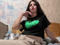 VickiLipoma with long dark hair, red lipstick, black shirt with green heart, beige pants, sitting on bed, hand on chin, confident, sexy, bedroom setting.