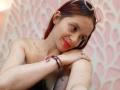 LesleyGray with red and brown hair, pink glasses on head, smiling, red lipstick, freckles, bare shoulders, colorful bracelet, arms resting on knees, soft pink and white patterned background.