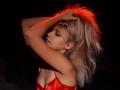 DataSley with blonde hair, arms raised, wearing a red satin top, red light on her hair, eyes closed, small tattoo on right arm, dark background, sensual pose.