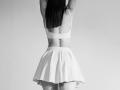 VirgieCrotts with arms up, back to camera, wearing a white sports bra and a flared skirt, showing a toned ass and long straight black hair. Black and white.