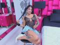 RoxyThunder with tattoos, black sheer lingerie, small cutout, blue skirt, white heels, sitting on chair, holding pigtails, pink and black room, smiling, looking at camera.