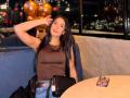 JennTylor with long black hair, brown sleeveless top, blue jeans, black handbag, and phone on table, sitting in restaurant, hand in hair, city lights in background.