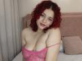 RosalieVittoria with red curly hair, pink lace bra, showing her big boobs, smiling, looking at camera, on pink bed, soft lighting, pale skin, pink lipstick.