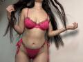 Tisia with long dark hair, wearing pink lace bra and panties with floral embroidery, holding her hair, showing her tits and pussy, standing against a plain wall.