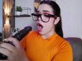 AryButty with black glasses, orange shirt, and pink lipstick, holding a dark dildo, mouth open in surprise. She has dark hair, freckles, and is looking at the dildo.