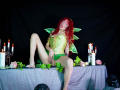GaiaDoll with red hair, green fairy wings, and yellow corset, sitting on a table with candles and flowers, spreading her green panties, showing her pussy, in a dark room.