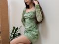 AmeliaMaiv with long brown hair wearing a green, wavy-patterned dress, showing her tits and ass, standing against a white wall with a potted plant on the left.