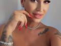 NicolSNaya with red nails, diamond necklace, and tattoos on her arm and chest, smiling at the camera, showing her boobs and a nose ring.