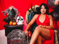 LilyConorr in a red dress, sitting on a throne with her legs crossed, wearing gold bracelets, with a skull, pumpkins, and a black crow on a table. Boobs, ass.