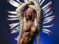 DaniaMarcell with pink hair, tan skin, and large tits, wearing a beige cardigan, feather headdress, and tattoo reading "Batty Girl" on her chest, arms raised.
