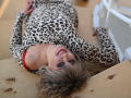 CandyLadyZ lying on floor, wearing leopard print dress, red bra visible, smiling, wavy blonde hair, makeup, hands near white furniture, wooden floor, relaxed, sexy, attractive.