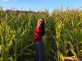 TrishMuhlbauer stands in a cornfield, sunlit, wearing a red off-shoulder top and gray pants, head tilted back, eyes closed, long dark hair, green cornstalks all around.