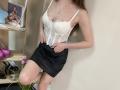 StellaNoir in a white lace bustier with boobs slightly visible, black mini-skirt, hands in pockets, long brown hair, necklace, standing against a wall, legs crossed.
