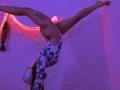 VioletaBlaze is upside down, wearing a white floral leotard, with legs spread, exposing her ass and a tattoo on her right thigh. Purple and pink lights illuminate.