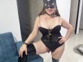 SamanthaGold with black lace corset, black mask, thigh-high boots, and beaded necklace, showing her tits and a pink butt plug, standing with one leg on a blue chair.