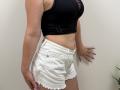 Tilliana with a black, cut-out, sleeveless top and white, frayed, denim shorts, showing her slim, tan, toned body and small, perky, bare tits. Hands on her hip.