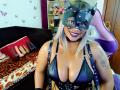 NashaJonny with cat mask, black leather bustier, and straps, showing big tits, smiling, blonde hair, tattoo on right arm, purple lipstick, in a purple gaming chair.