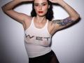 Venusinylons with wet tank top, white with "WET" text, showing her tits, black panties, tattooed arm, red lipstick, and dark hair, hands in hair, standing, serious expression.