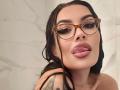 ZoeyMarie with wet black hair, tortoiseshell glasses, and her tongue out, showing glossy pink lips, tattoo on her left shoulder, and a necklace.