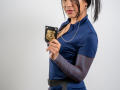 VelvetRoss with dark hair in a ponytail, wearing a blue police shirt, holding a police badge, has a necklace and arm bands, standing, looking at the camera.