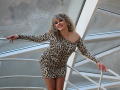 CandyLadyZ with curly blonde hair, wearing a leopard print dress, smiling, leaning on a white railing, showing off her ass in a tight, off-shoulder, long-sleeve mini dress.