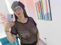 MadameScarlett with glasses, black mesh top showing her tits, beanie, and red lipstick, taking a selfie in a room with abstract art. Holding a silver iPhone.