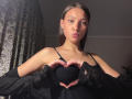 KassandraDalal with pouty lips, making a heart shape with her hands over her black bra-covered boobs, wearing a black lace top, and a thin necklace, in a dim room.