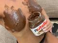 SexxyGirl's chocolate-covered tits are in the foreground with a jar of Nutella in her hand, some chocolate on her fingers, and a beige textured couch in the background.