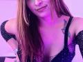 AnaEox with long brown hair, black sparkly dress, low-cut showing tits, tattoo of bird on right chest, purple light, subtle smirk, nose ring, wrist bracelet, slightly bent forward.