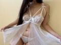 Tisia in white lace lingerie with sheer babydoll, showing her tits in a bra with bows, holding the fabric, long dark hair, beige wall, chain necklace, white panties.