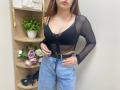 Rioria with her boobs in a sheer black top, blue jeans, and a belt, standing in front of a wooden shelf with flowers and high heels.