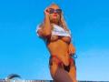 MademoiselleBella with blonde hair, sunglasses, white shirt lifted to expose her tits, black thong, and a sun-kissed tan against a bright blue sky. Her ass is toned.