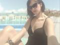 MiraMixi with short black hair, wearing dark sunglasses, black textured top, and colorful bracelets, sitting by a pool, showing her boobs and a cute, relaxed face.