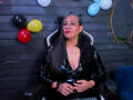 VictoriaKiley in a black shiny latex zip-up top, sitting in a chair, hands on her belly, long black hair, red lipstick, gray wood wall, balloons in background.