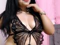Zaria69 with long black hair, wearing a sheer black lace butterfly bra, exposing her tits, a pearl necklace, and a gold watch, against a pink curtain.
