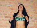 AnneStone with green lace bra and strappy underwear, black jacket open, showing tits, tan skin, long dark hair, eyes closed, necklace, standing against brick wall.