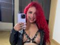 Estefy69 with red curly hair, smiling, taking a mirror selfie with a purple phone, wearing a black leather harness that exposes her tits, and a black jacket.